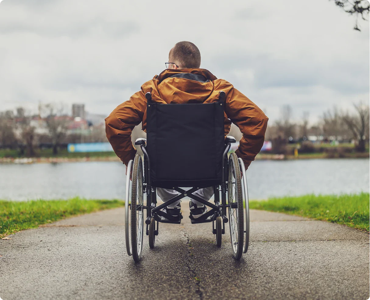 A person in a wheelchair, wearing a brown jacket, facing a tranquil lake, with cloudy skies and greenery lining the pathway.