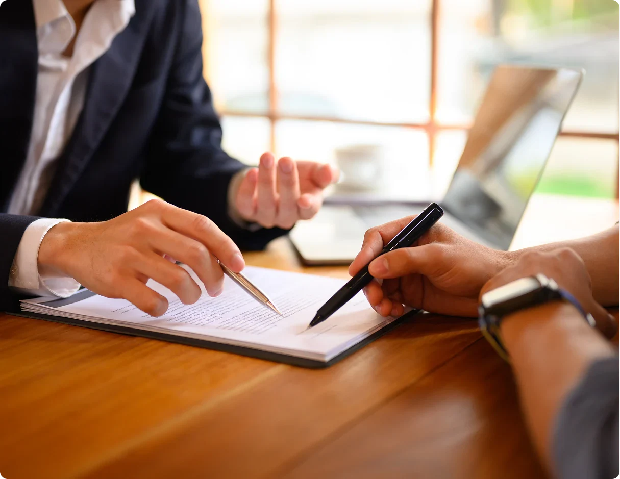 Two people engaged in a business discussion, one pointing at documents while the other writes notes, with a laptop in the background.