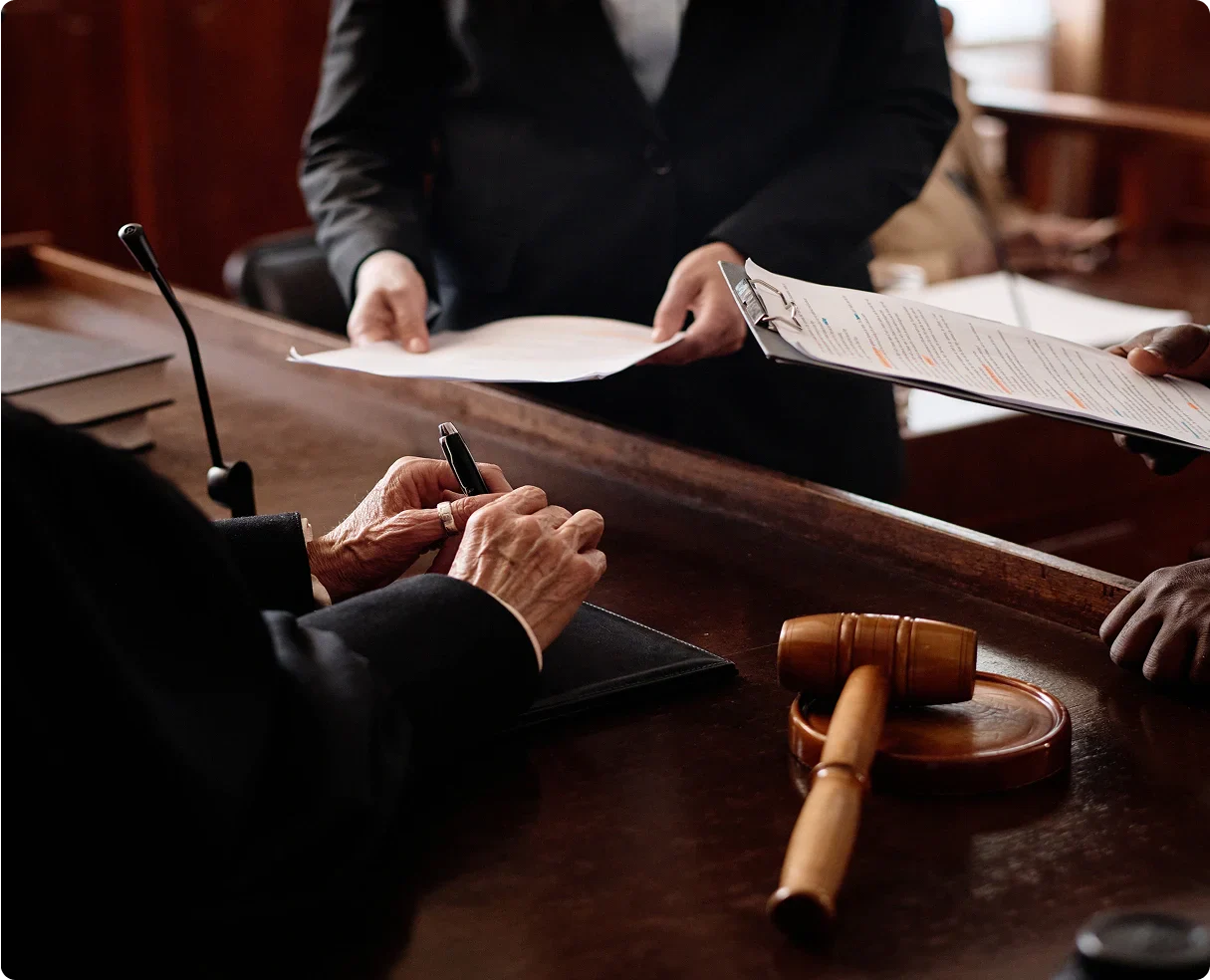 A judge's hands with a pen near a gavel as a lawyer presents documents in a courtroom setting.